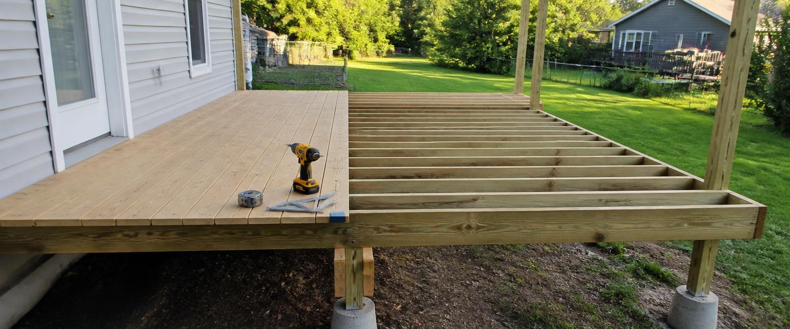 Backyard deck under construction with pressure-treated joists at 16 inch centres, deck boards being laid with a spacer, and concrete footings under the posts